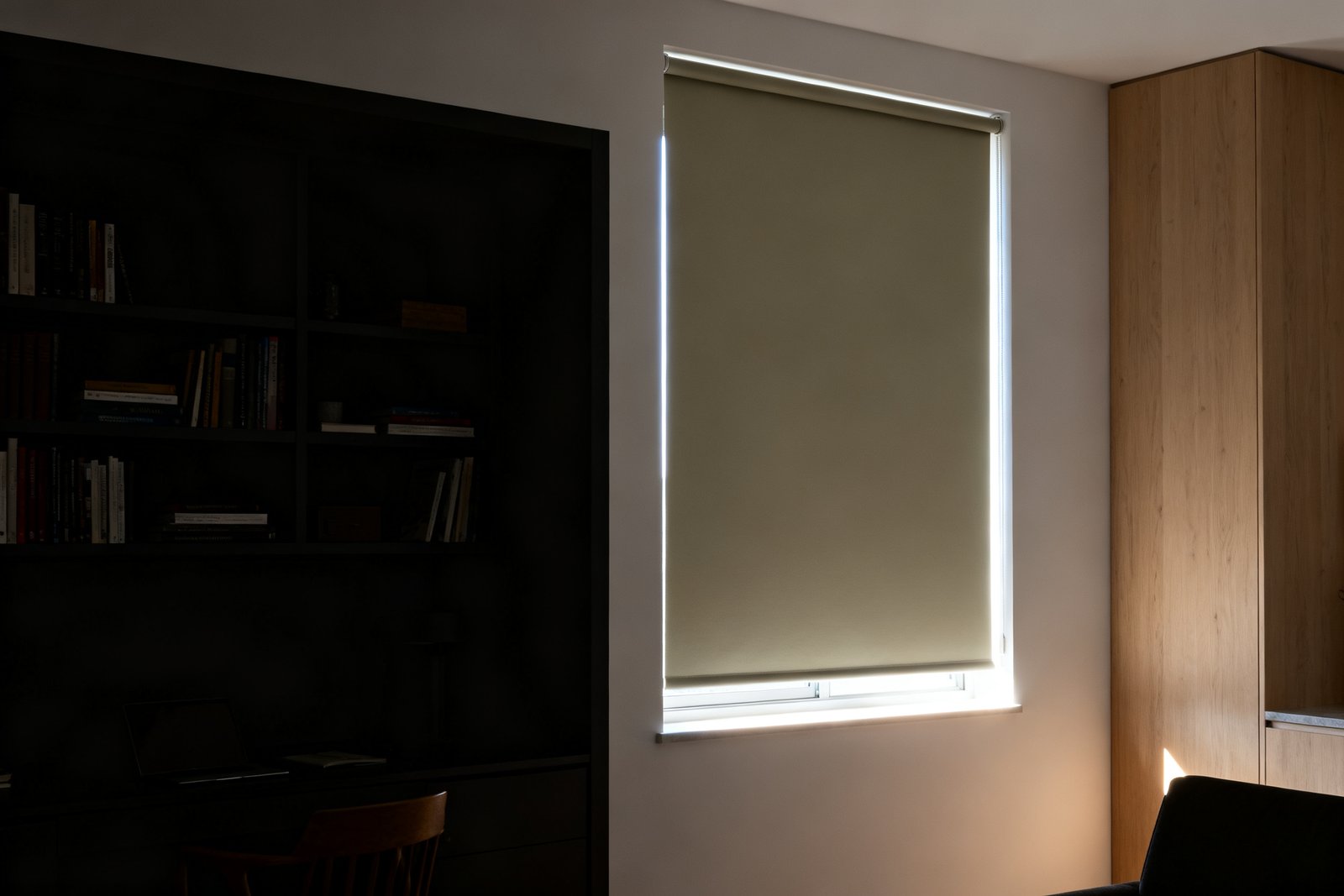 A blackout roller shade fully lowered in a darkened study, thin lines of daylight leaking down both side gaps and under the bottom bar.
