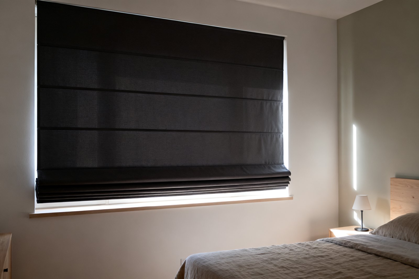 A charcoal blackout roman shade fully lowered in a dim bedroom, its horizontal folds visible and daylight tracing the side and bottom gaps.
