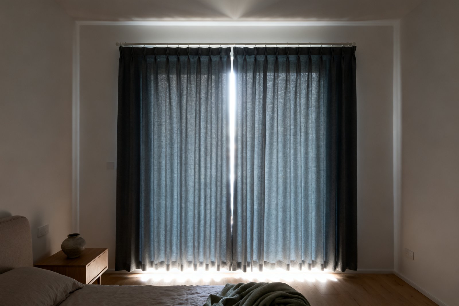 A dim bedroom at midday with a room-darkening curtain drawn, daylight escaping in a bright halo around the top, sides and hem.