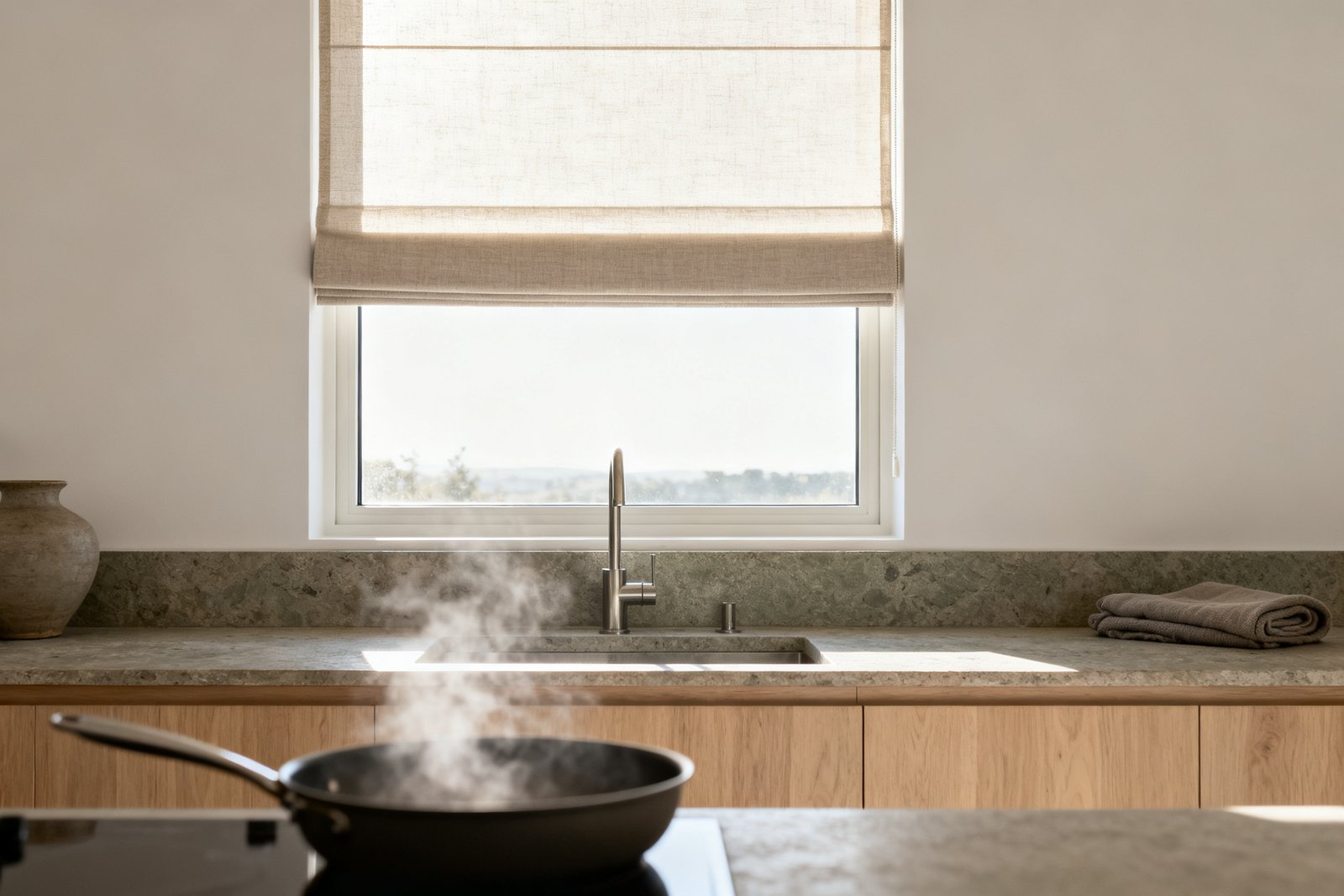 View along a kitchen worktop to the window above the sink, a short roller shade half down, a pan steaming in front